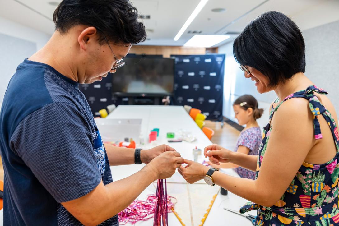 Two standing people work together on a craft project, with young workshop participants in the background