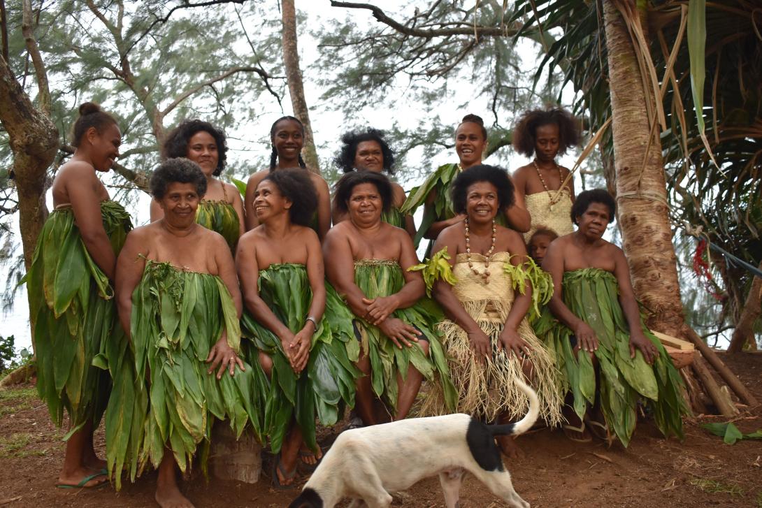 A group of women, clothed in dresses made from plant materials, with a dog in the foreground