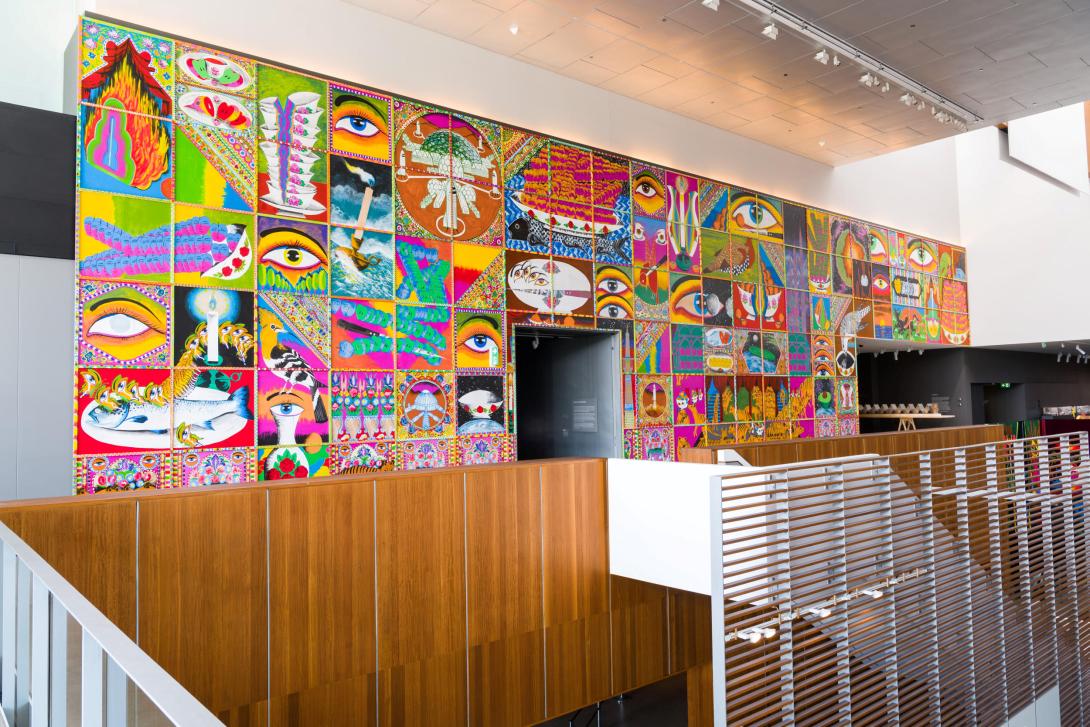 An installation view of a large, colourful mural installed in a gallery space overlooking a staircase