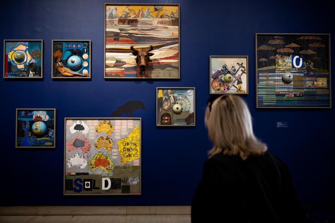 Oil paintings installed salon-style on a dark blue gallery wall, with a person looking on