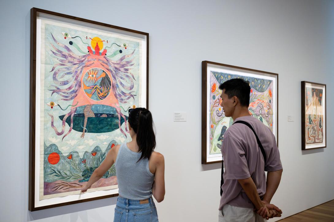 An installation view of three colourful works on paper in a bright gallery space, where two visitors look on