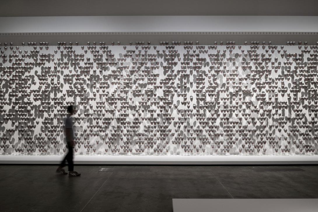 An installation of small metal skulls forming a curtain-like feature across a wide, white gallery wall