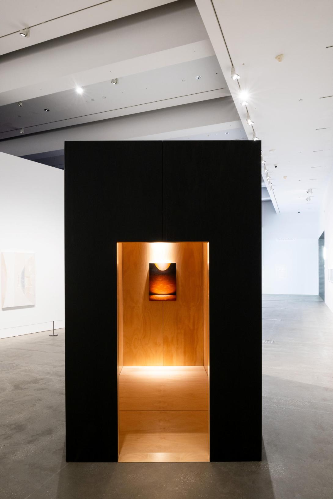 A black architectural form with a glowing doorway through which a painting can be seen; this whole form is installed in a brightly lit gallery space