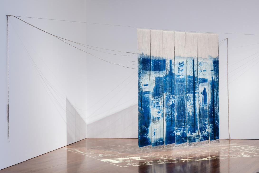 An installation view of a blue and white textile work installed suspended in a bright gallery space
