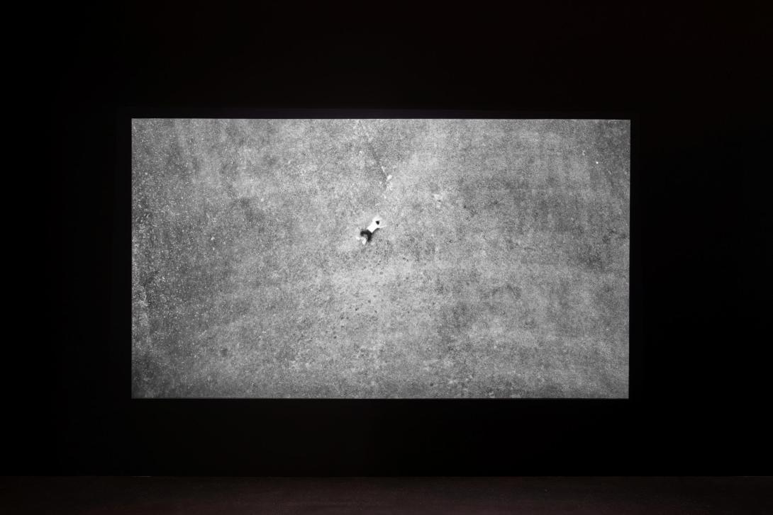 A black-and-white video still, seen installed in a dark gallery space