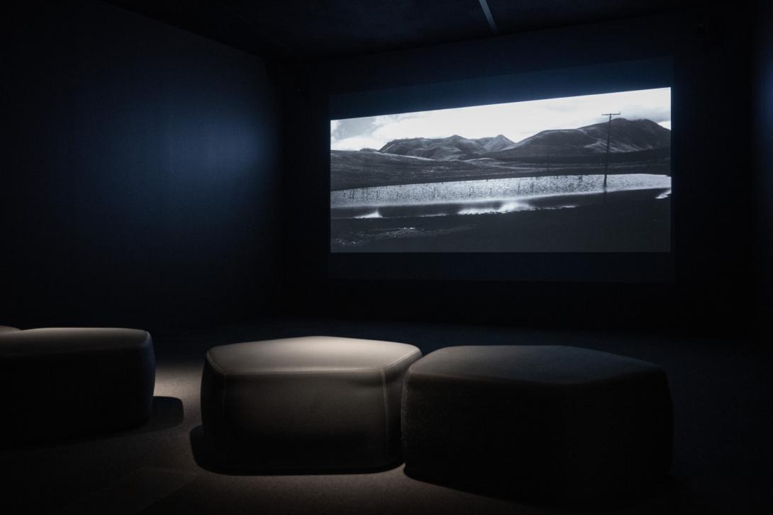 A black-and-white video being screened in a darkened gallery space, with grey seating