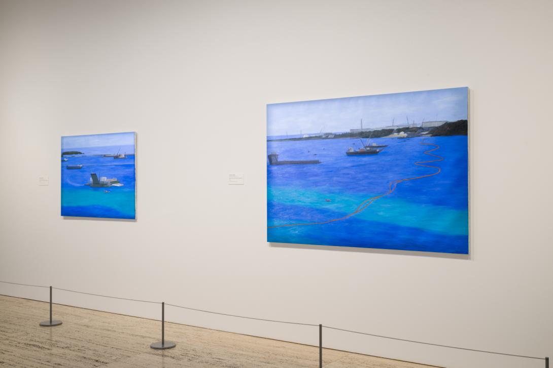 Two oil paintings depicting blue oceans with military vessels installed on a white gallery wall