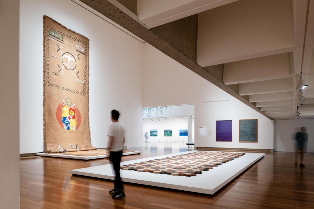 A large textile work installed in a gallery space with high ceilings, with a sculptural work made of tiles in the foreground; a visitor looks on.