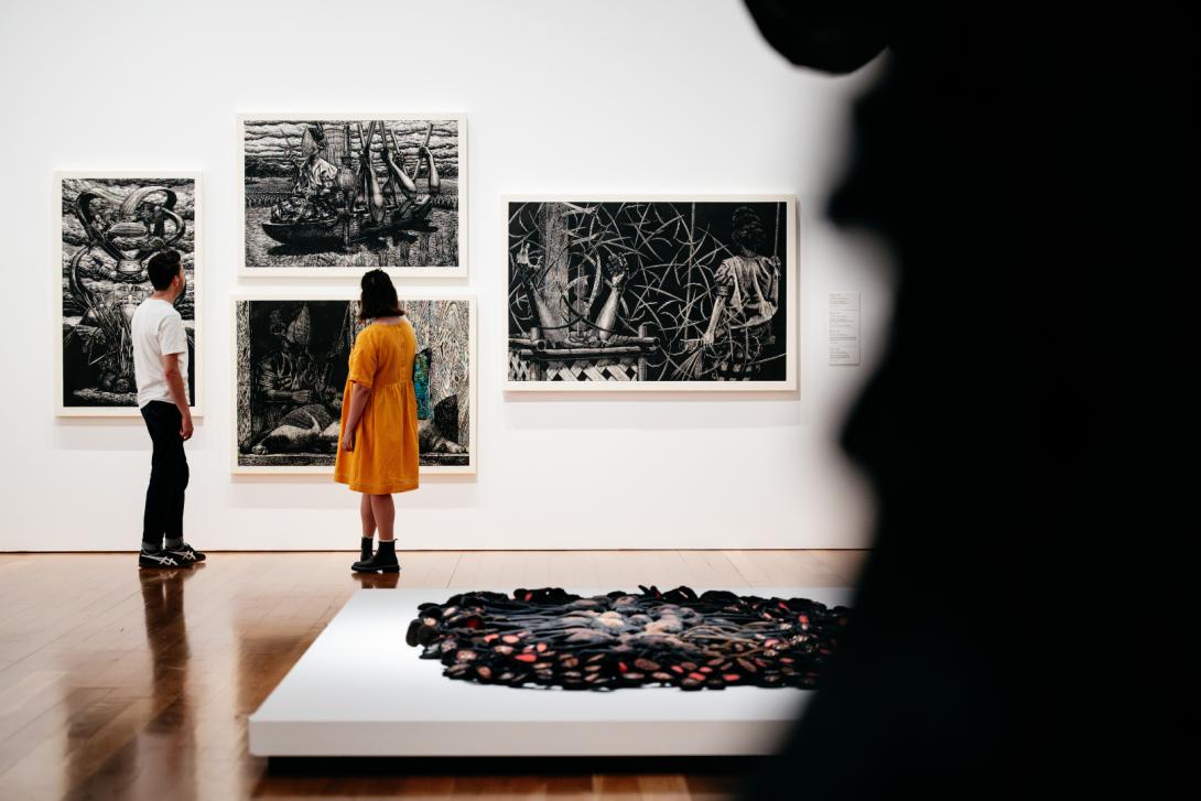 Two gallery visitors gaze at black-and-white woodcut works on a far gallery wall, with our view of the gallery itself obscured by woven sculptures in the foreground