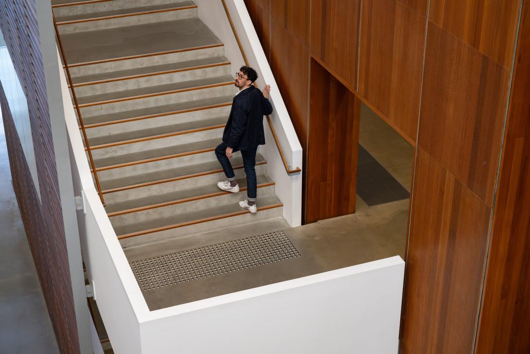 A man wearing a black jacket, jeans and white sneakers climbs the stairs in a gallery space while appearing to listen closely to an announcement or other broadcast audio