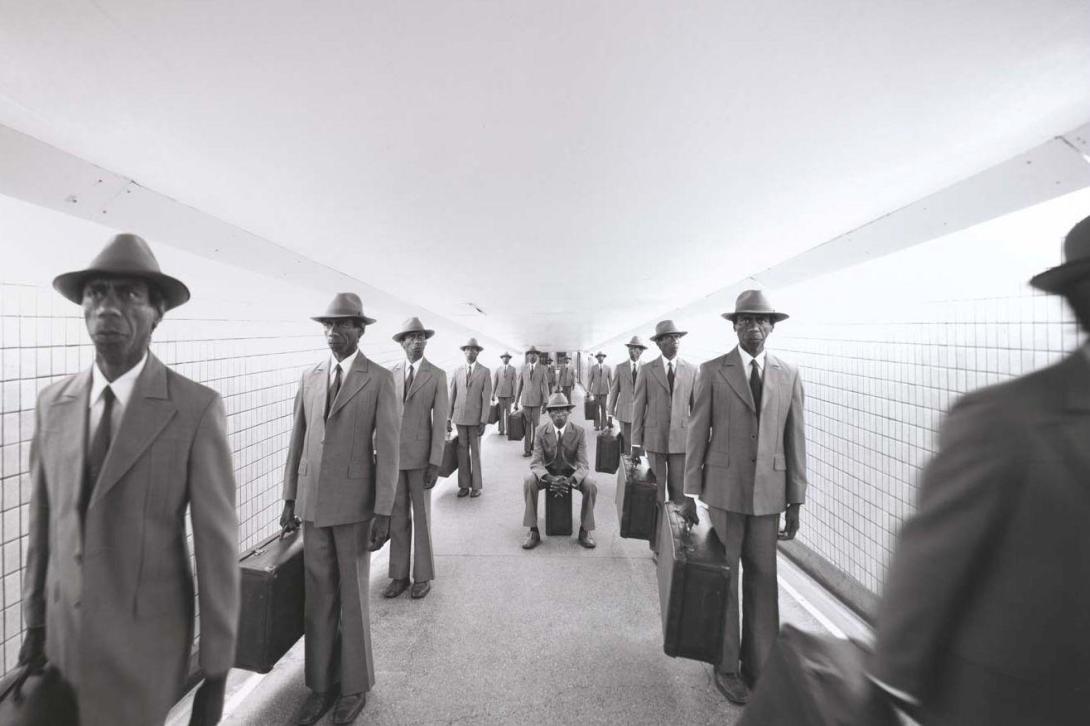 A black-and-white photograph in which duplicates of an Australian Indigenous man in a vintage business suit and hat stand in various commuter-coded poses in a tunnel