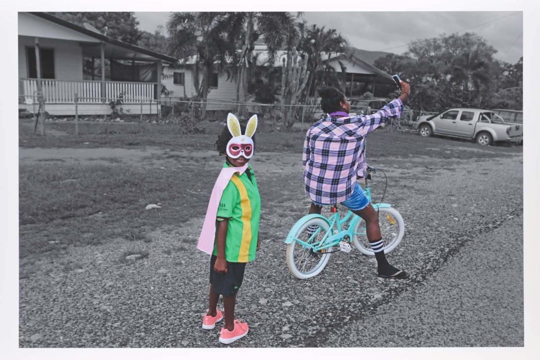 A detail view of a photograph of two children by Naomi Hobson: one rides a bike and faces away from the camera; the other child, wearing a cape and bunny ears, looks into the camera lens