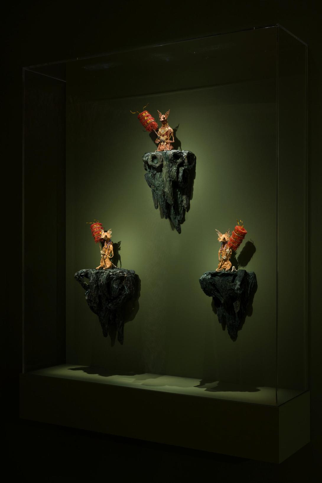 Three small puppets, made from joss paper, resembling deer-mice and foxes, installed on a dark green wall in a dim gallery space.