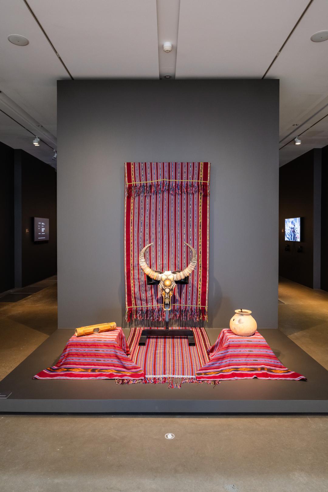 A view of a red textile installation, featuring a buffalo skull at centre, installed in a gallery space with dark grey walls.