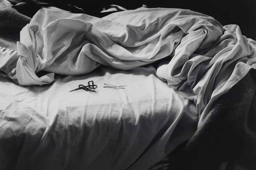 This portrait-oriented, black-and-white photograph depicts an unmade bed, with its white sheets rumpled. Two small tangled, wiry objects are visible on the flat on the fitted sheet.