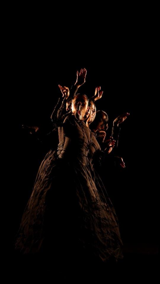 In this still from a portrait-oriented video work, a Sāmoan woman dressed in late nineteenth-century, western-style clothing is captured mid-dance. Illuminated against a dark background, a transparent motion trail of the figure's previous movements is also visible.