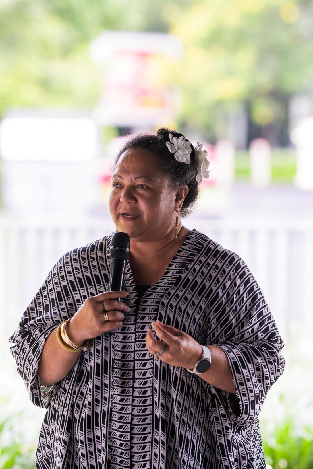 A photograph of an artist, Mele Kahalepuna Chun, holding a microphone and speaking to an audience. Green trees can be seen in the distant background.