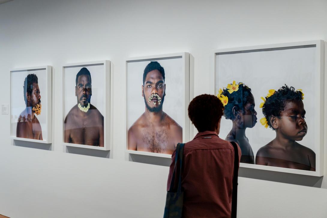 A photograph of a visitor walking past an installation of photographs on a white gallery wall; the photographs depict portraits of Indigenous Australian men adorned with flowers.
