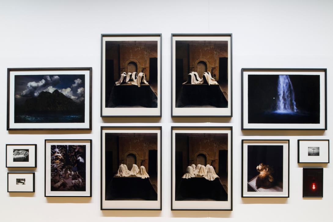 This photograph captures a view of a salon-style installation of photographs, with very small works hung at the far edges and the largest works hung in the middle. Multiple artists are represented; however, each photograph included has dramatic, Baroque elements in its composition.