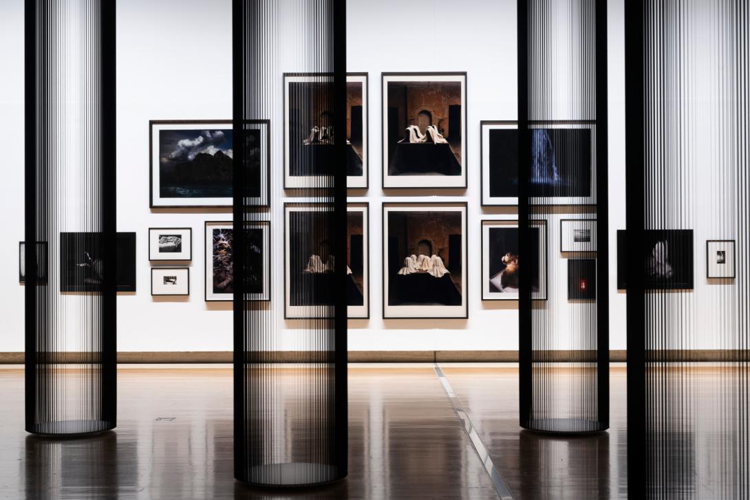 This photograph captures a view of a salon-style installation of photographs, with very small works hung at the far edges and the largest works hung in the middle. In the foreground, columns made of tape from inside VHS cassettes appear both monumental and transparent. Multiple artists are represented; however, each photograph included has dramatic, Baroque elements in its composition.