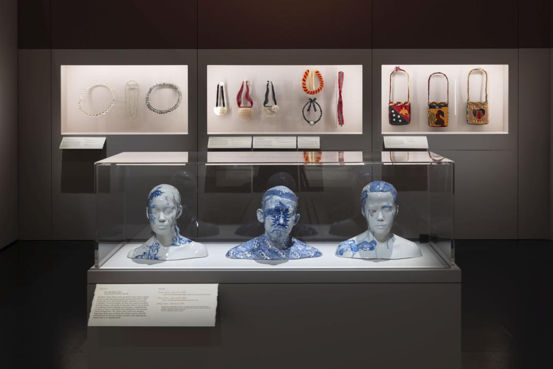 In a gallery space, three porcelain busts are installed in a glass vitrine in the foreground while, in the background, adornments including necklaces and bags are installed in a backlit glass case along the back wall.