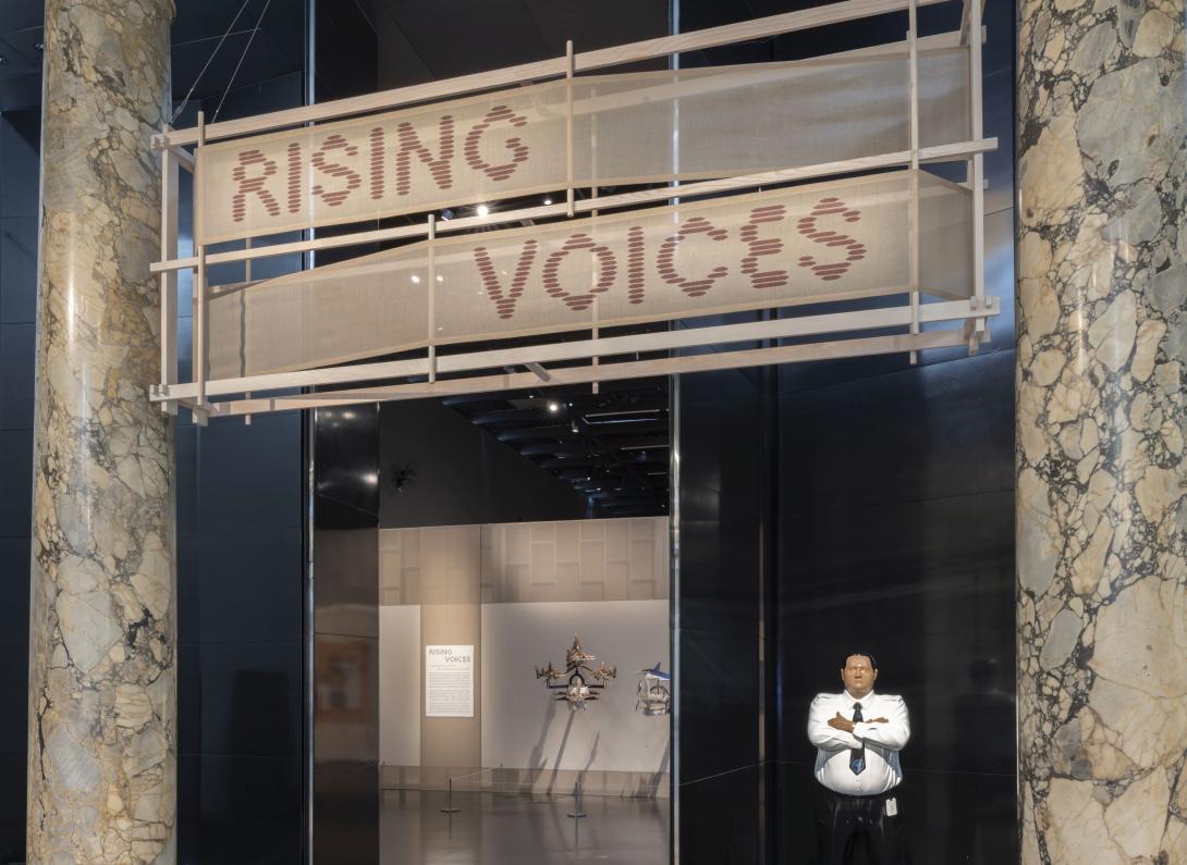 A view of the entrance of an exhibition called 'Rising Voices', with marble columns either side of the title banner, and with a sculpture of a security guard visible at the exhibition entrance.