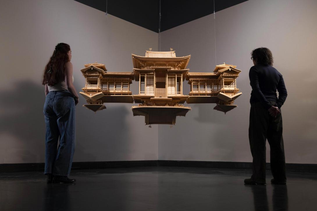 An installation view of a wooden sculpture taking the form of a Japanese temple, doubled as if in reflection; two people view the work