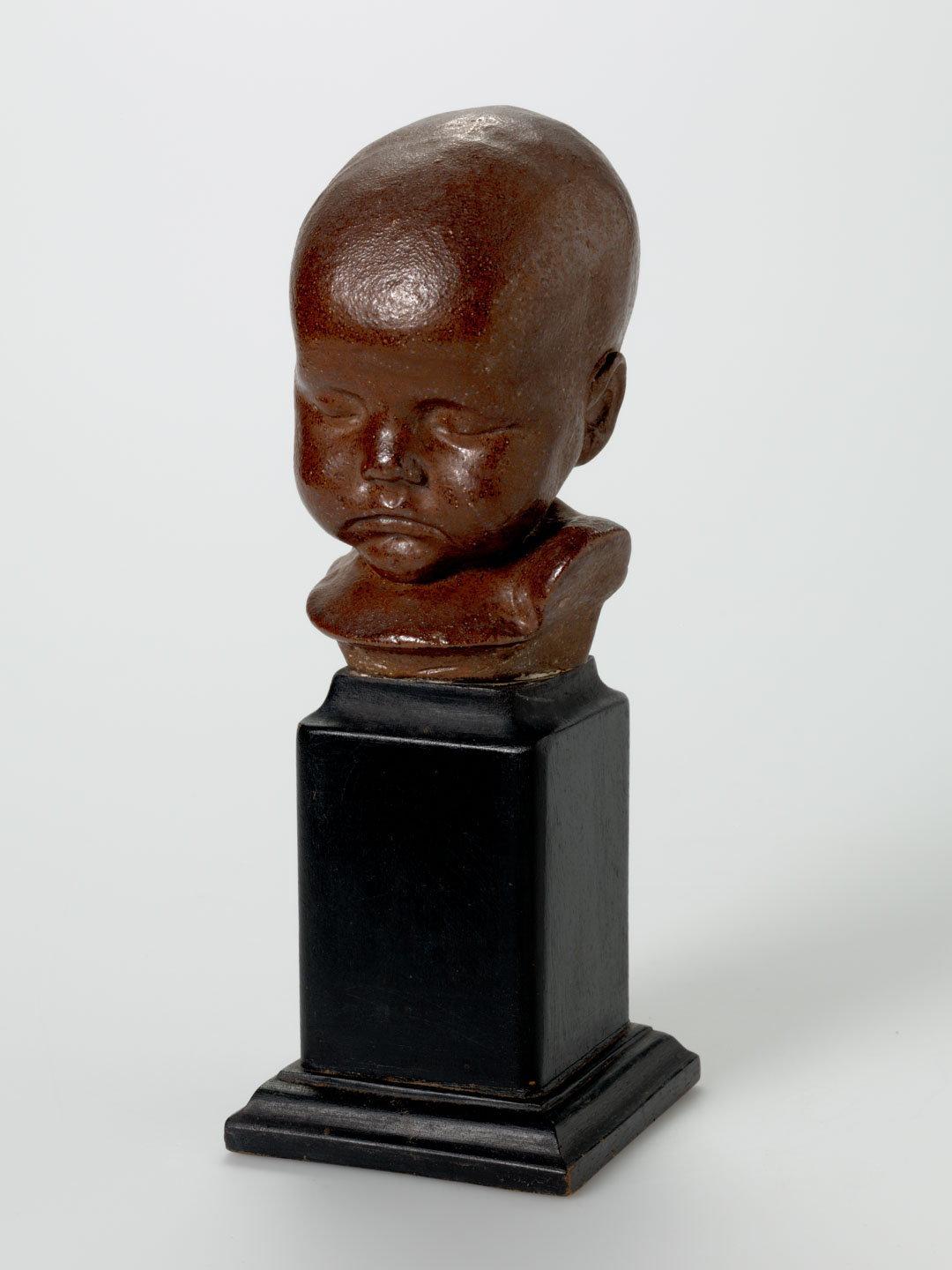 Artwork Sculpture:  (Baby's head) this artwork made of Stoneware, Darra pipe clay modelled and salt glazed with wooden base, created in 1935-01-01