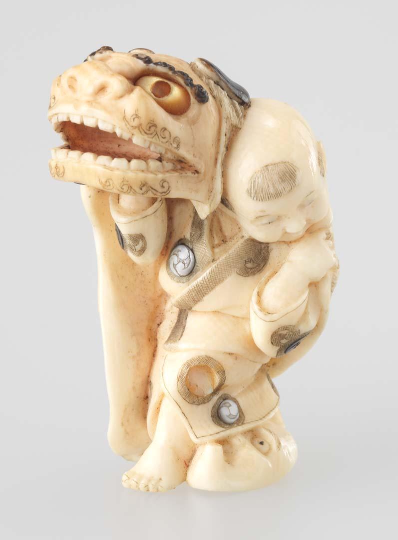 Artwork Netsuke:  (child with a large monster mask) this artwork made of Carved ivory with inset mother of pearl and red lacquer with painted details, created in 1800-01-01