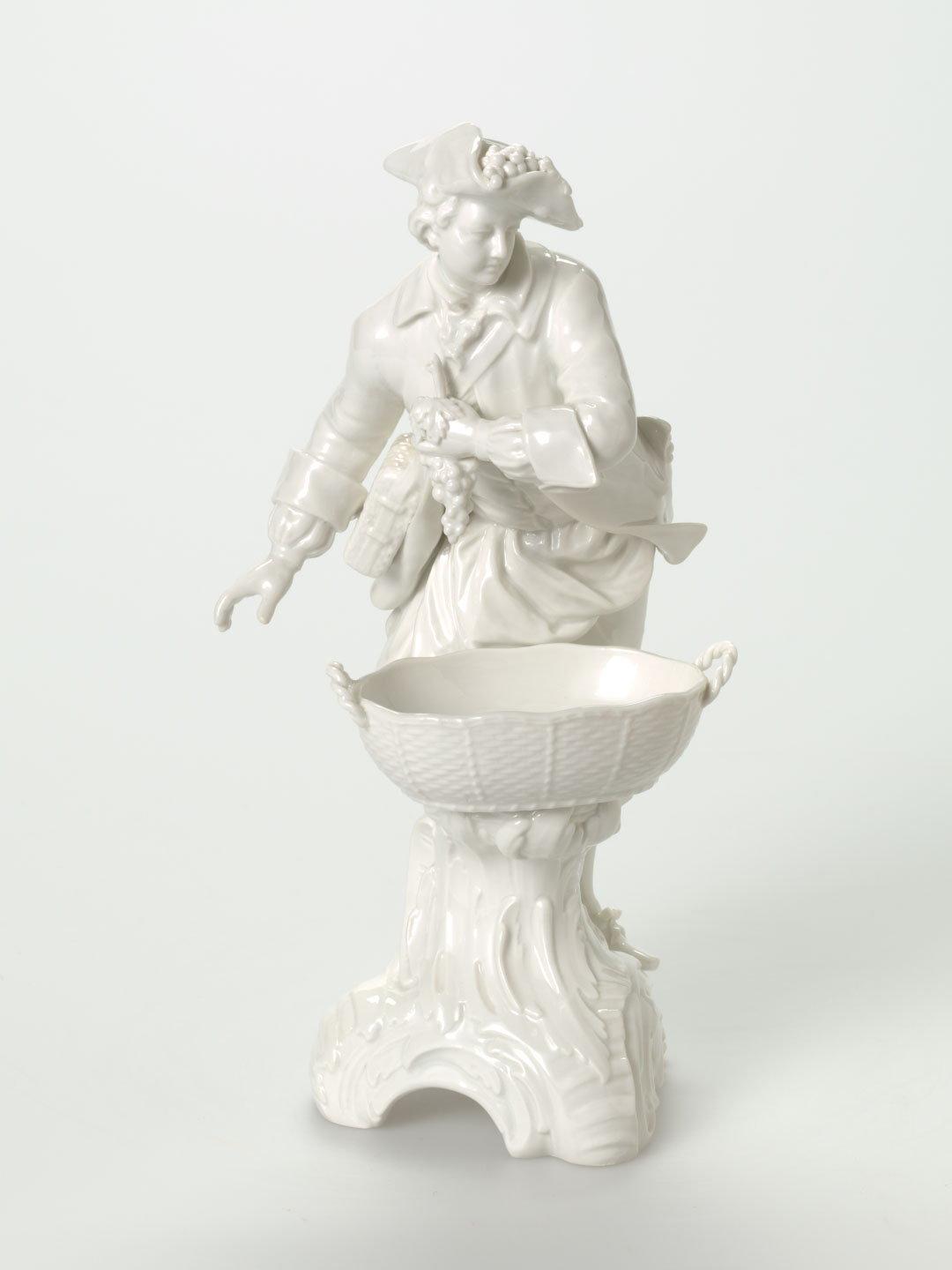 Artwork Figurine:  (gallant holding a bunch of grapes) this artwork made of Hard-paste porcelain finely modelled figure of a gallant holding a bunch of grapes over a basket, Rococo base and clear glaze, created in 1870-01-01