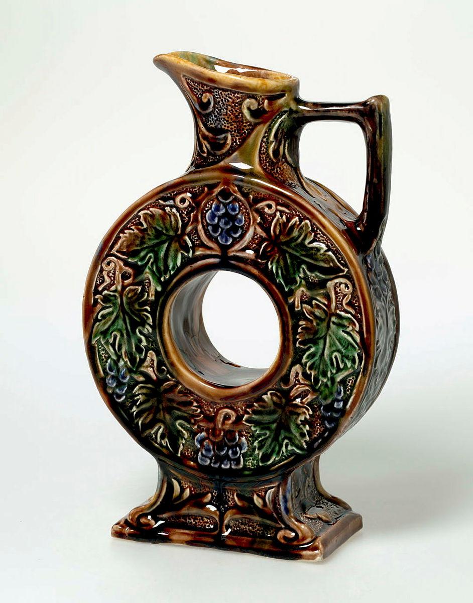 Artwork Wine jug this artwork made of Earthenware slab and hand-built pierced circular flask (pierced with a hole) on a stepped stand, carved with grapes and leaves and glazed brown and colours, created in 1930-01-01