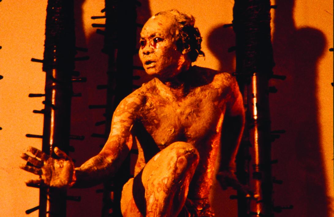 A photograph of an artist during a performance; wearing body paint, he crouches in the orange light of a gallery space.