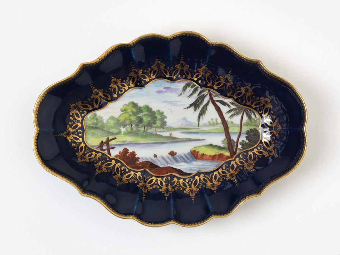 Artwork Lozenge-shaped dish:  (river landscape) this artwork made of Soft-paste porcelain, slip-cast scrolled form with deep blue border with river scene in green, blue, brown and purple.  Gilt dentil edge and rich gilt border with greenish glaze, created in 1768-01-01