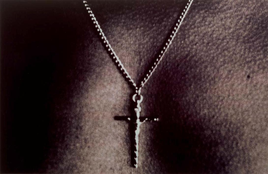 A close-up photograph of a masculine chest with a metal crucifix worn on a chain necklace