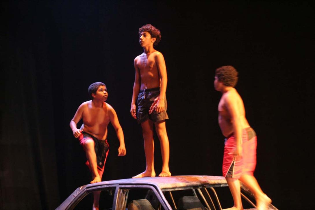 A photograph of three young Indigenous boys climbing on the roof of a car during a performance by Bangarra Dance Theatre.