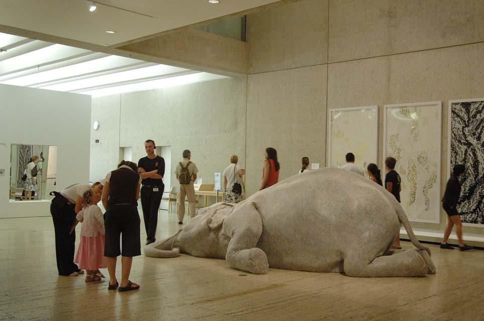 An installation view of a life-size and very realistic sculpture of an elephant, slumped to the floor as if dead, in a gallery space surrounded by onlookers.