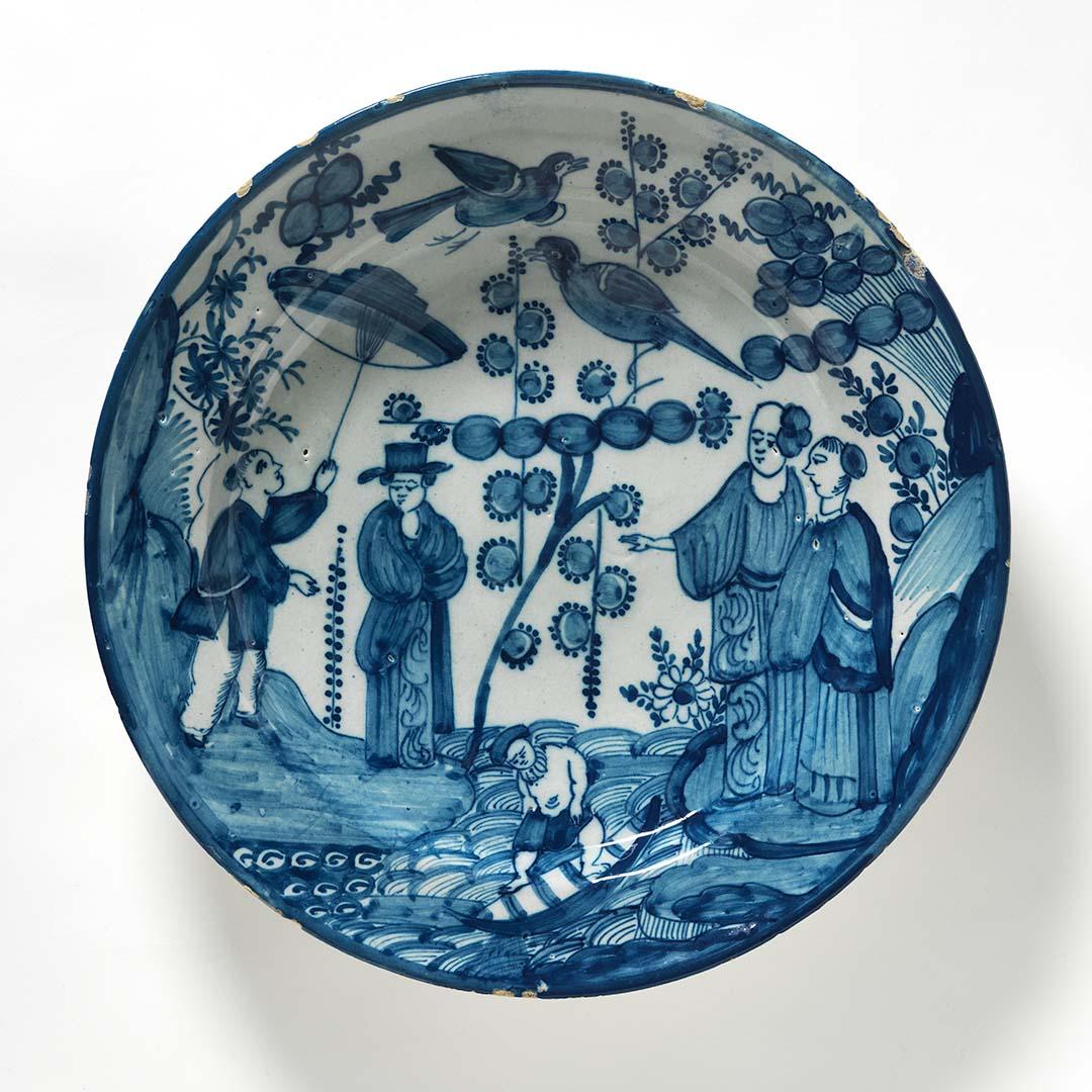 Artwork Charger, decorated with blue and white Kakiemon design showing a man with a boy attendant with parasol and two women looking across a river, a fisherman in a boat and two birds flying overhead this artwork made of Earthenware, wheelthrown and tin glazed with cobalt decoration