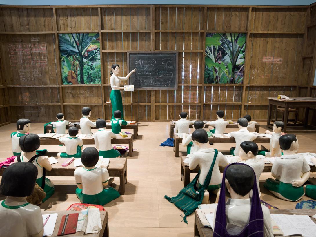 A close-up installation view of a sculptural work depicting a Myanmar school classroom with children seated at low benches, and with a teacher at a blackboard.