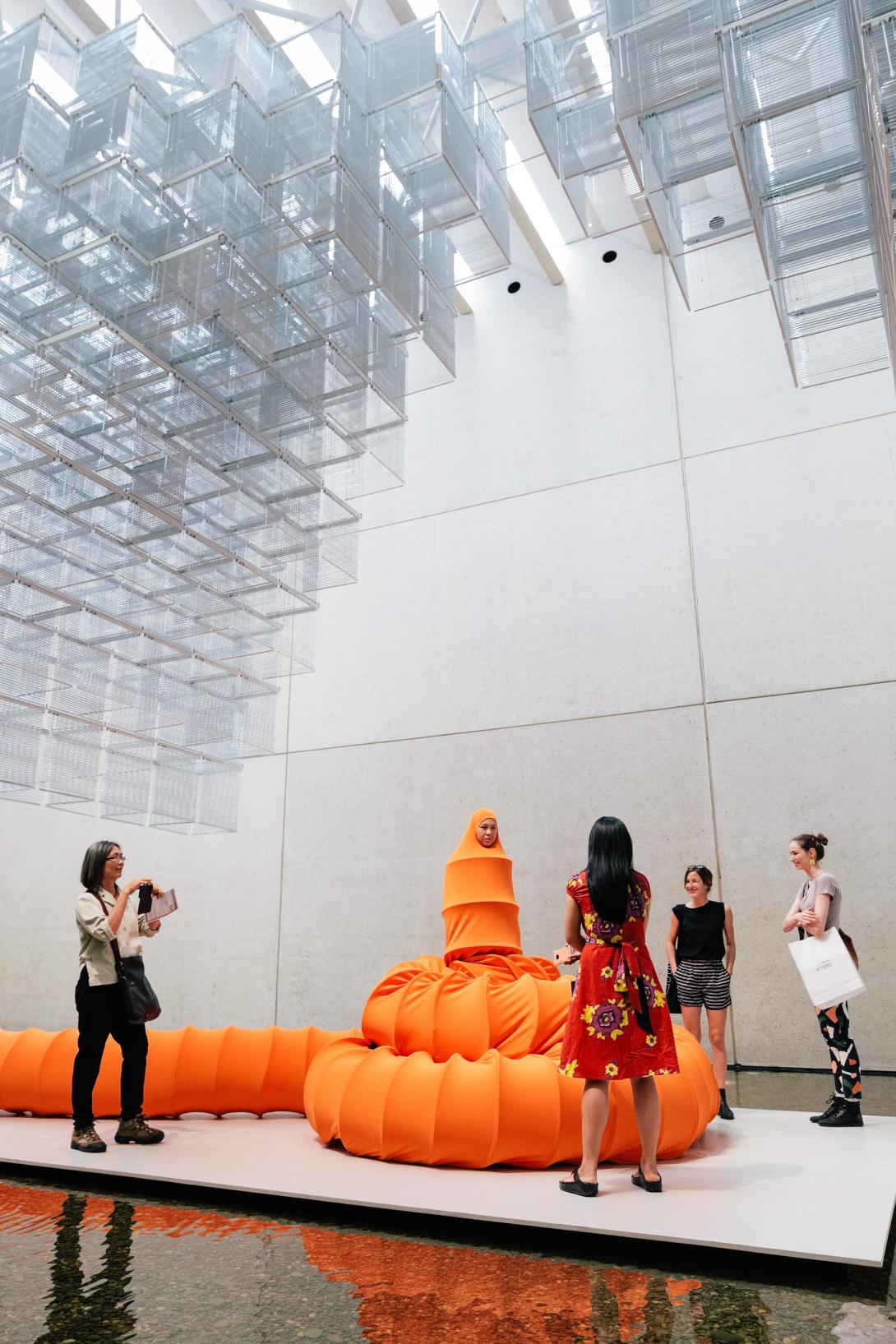A view of a performance in which an artist dressed as an orange caterpillar performs under a suspended installation.