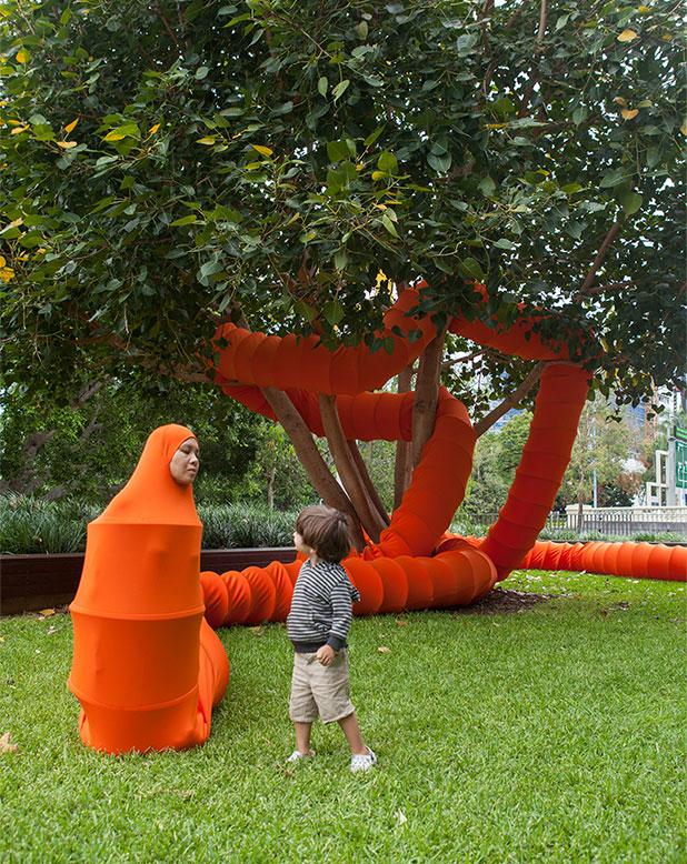 An artist dressed as a red caterpillar, with her body and tail threaded around a tree on the QAGOMA grounds, meets a little boy visiting the Gallery. 
