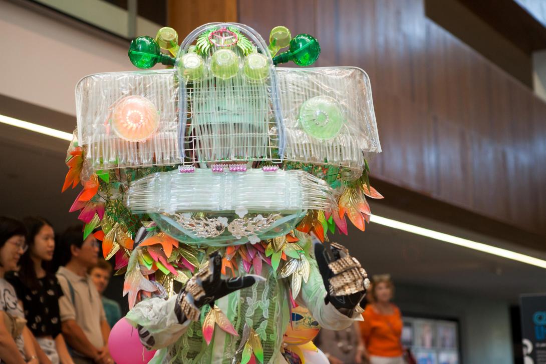 A dancer costumed as a mythical creature with huge square eyes lit orange and green stands in the GOMA foyer space.