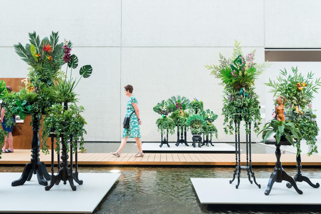 An installation view of a visitor walking through QAG's Watermall, on a path over the water bordered by plants on stands.