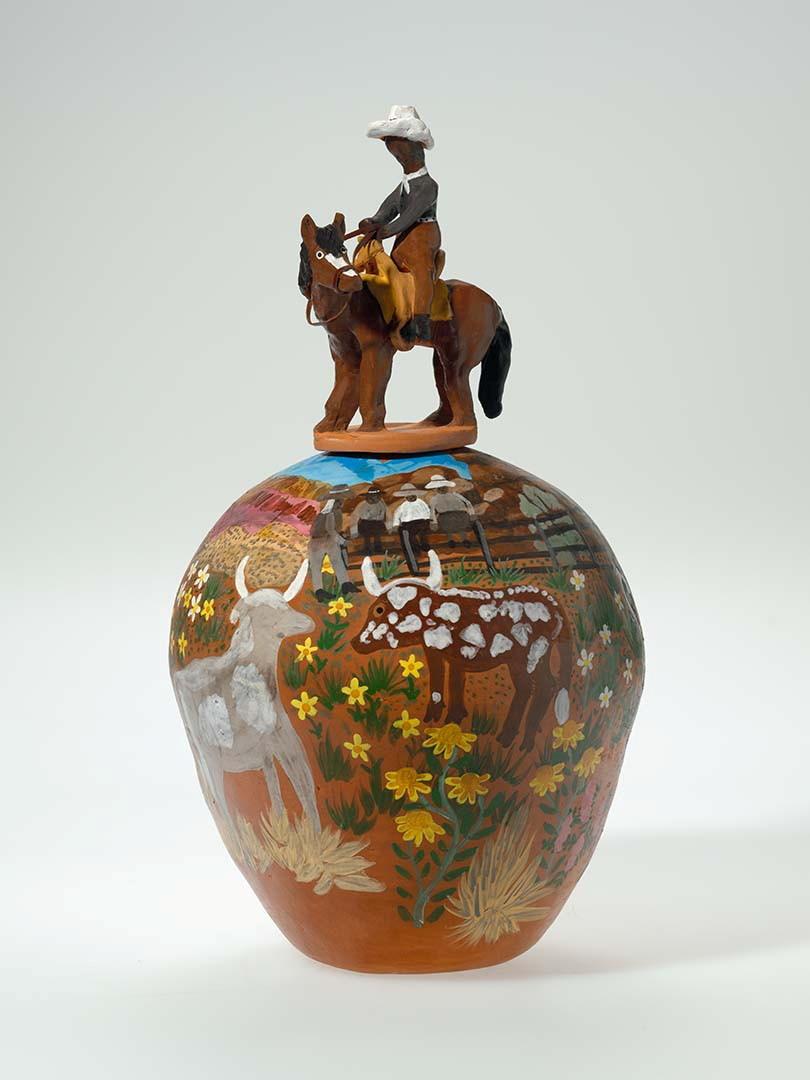 An earthware pot painted with a farm scene, with a sculpture of Albert Namatjira riding a horse on top as the lid of the pot.