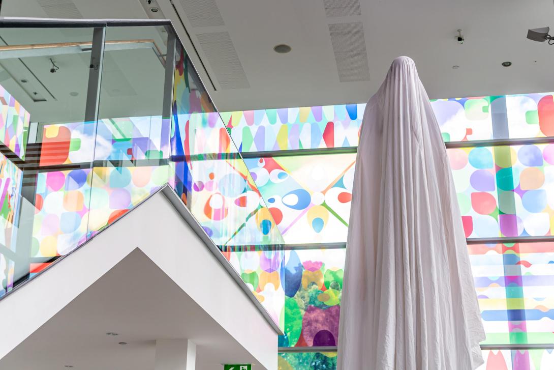 Completely cloaked in a white sheet, performance artist Brian Fuata stands before Shannon Novak's colourful APT10 window installation in the bright light of GOMA's Long Gallery.
