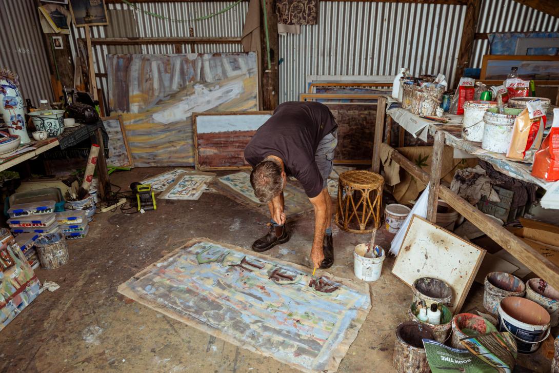In his shed-studio, an artist bends down to work on his artwork, which is laid flat on the floor; around him are chaotic shelves and piles of paints and materials.