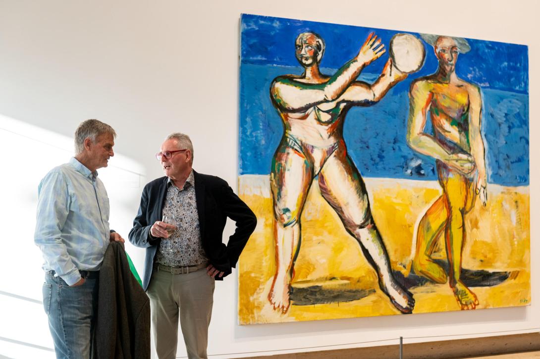 A photograph of two men in business casual standing to the left of a large, bright work of art installed in a gallery; the work depicts two naked people playing volleyball on a beach.
