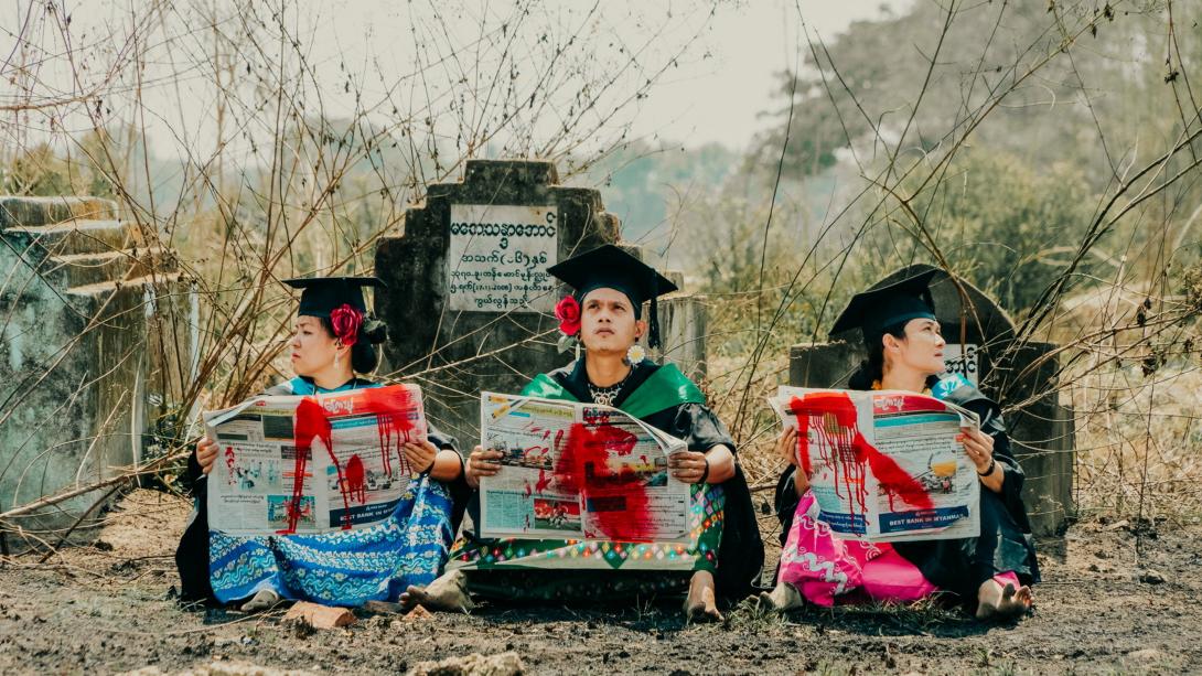 Three graduate students wearing mortarboards sit reading blood-splattered newspapers in a cemetery in Myanmar.