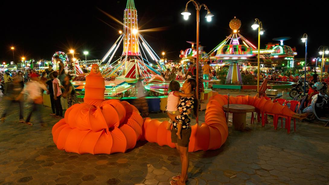 A still photograph taken at night in a fun fair; the artist is costumed as a red caterpillar-like bug with a very long body and tail.