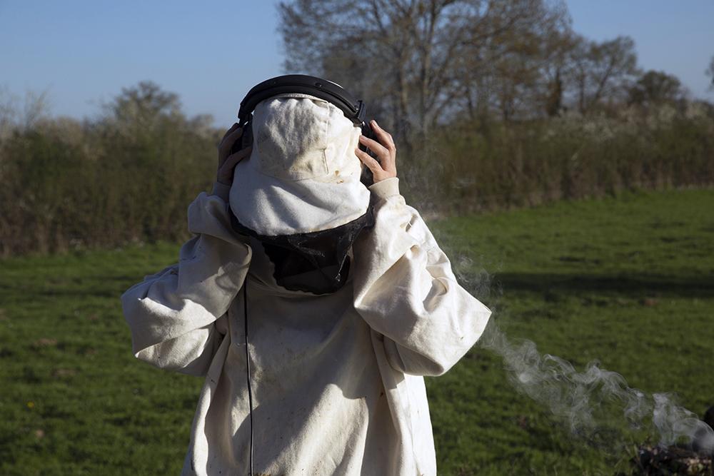 A person dressed in white protective clothing stands in a field with headphones on, recording the sound of a beehive.