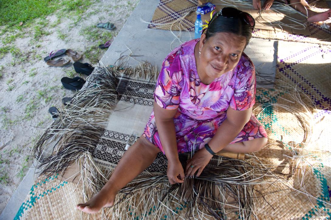 A woman sits on the ground weaving a mat; she is weaving a purple dress and looking up at the camera, smiling.